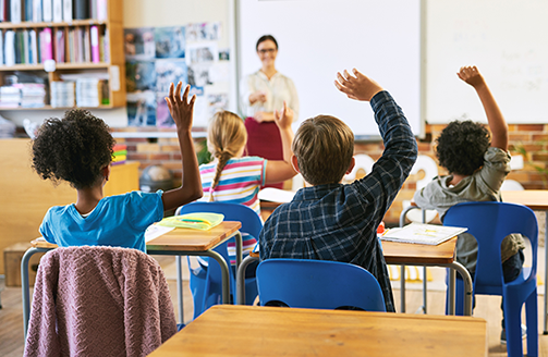 A female teacher facing four students with raised hands in a classroom.