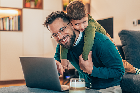 A father looking at a laptop computer on a table while his young son hangs on his back, looking over his shoulder.